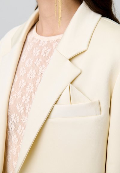 Blazer couleur crème avec des revers crantés, tissu texturé et une pochette pliée. En dessous, un haut en dentelle avec des motifs floraux.