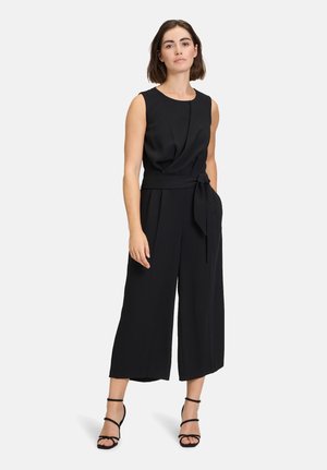 Tuta nera senza maniche con una vita aderente fissata da un nastro, pantaloni a gamba larga e una texture liscia. Design semplice senza motivi o decorazioni.