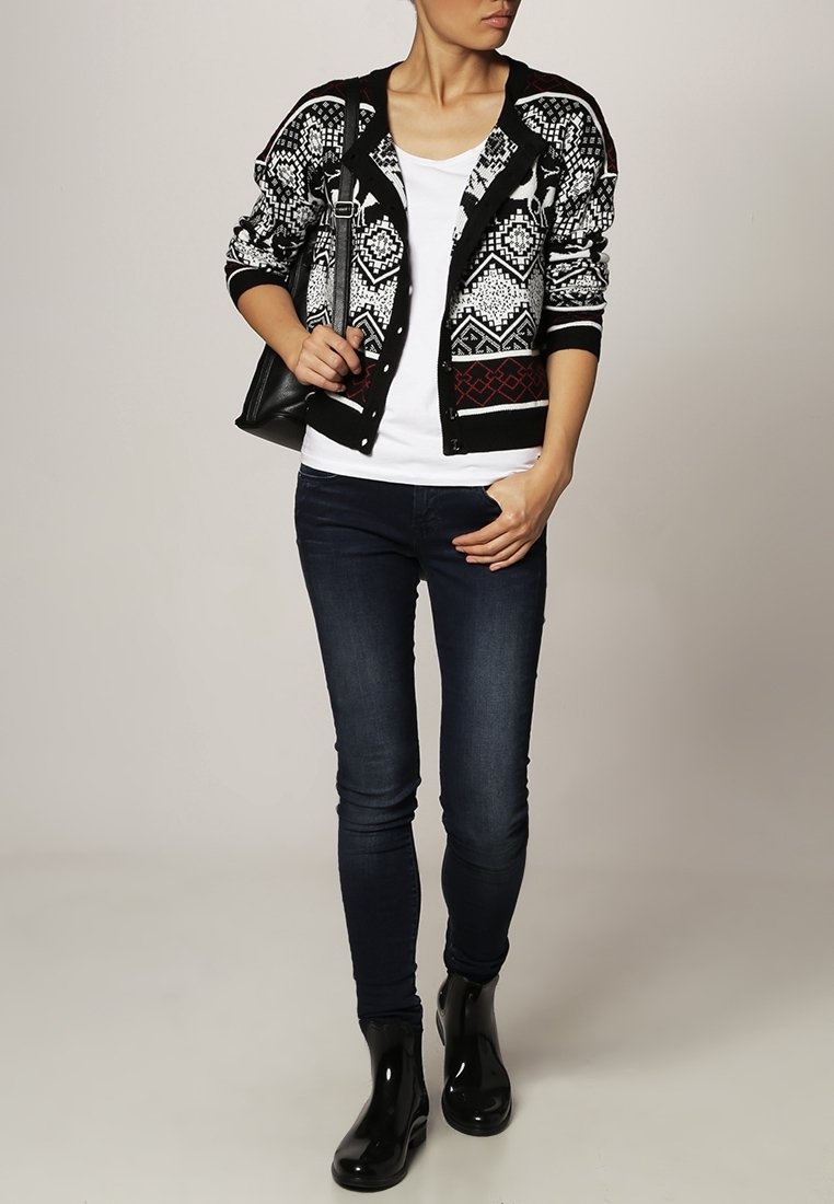 Femme portant un cardigan à motif noir et blanc, un t-shirt blanc, un jean skinny foncé, des bottes noires et un sac à bandoulière noir.