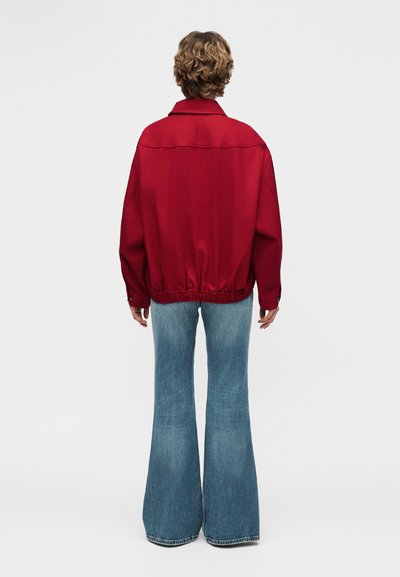 Άτομο με κοντά σγουρά μαλλιά που φοράει κόκκινο μακρυμάνικο μπουφάν και μπλε τζιν με φαρδύ καμπάνα, στέκεται πλάτη σε ένα απλό φόντο.