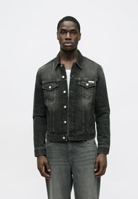 Čierna džínsová bunda s gombíkmi vpredu, dvoma náprsnými vreckami a viditeľným prešívaním. Hladká textúra s priliehavým strihom, doplnená sivými džínsami.