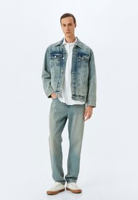 Giacca di jeans con finitura blu rovinata, due tasche sul petto, abbinata a jeans blu chiaro e una camicia bianca, completata da sneaker bianche.