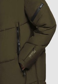Giacca imbottita verde con tasche con zip e finitura testurizzata. Presenta un logo visibile sulla manica e dettagli in zip neri.