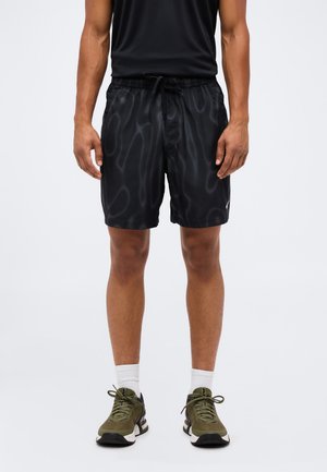 Erwachsener trägt schwarze, gemusterte Sportshorts, weiße Socken und olivgrüne Turnschuhe und steht vor einem schlichten Hintergrund.