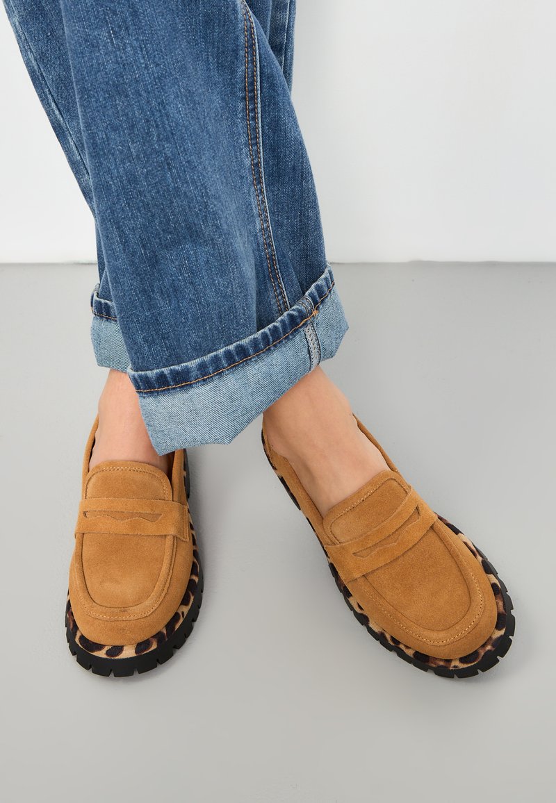 Zamszowe loafers w kolorze brązowym z czarną i w panterkę podeszwą; zaokrąglony nos i haftowane akcenty. W zestawie z podwiniętymi niebieskimi dżinsami.