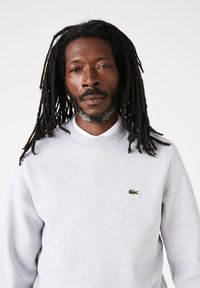 Grijze katoenen sweatshirt met een ronde hals, voorzien van een klein groen krokodillenlogo op de linkerborst. De stof lijkt zacht en textuurrijk te zijn.