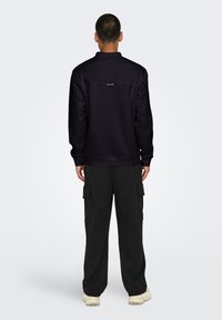 Schwarze Zip-Up-Trainingsjacke mit minimalistischem Design, runder Kragen und Logo-Detail auf dem Rücken. Kombiniert mit schwarzen Cargo-Hosen und hellen Sneakern.