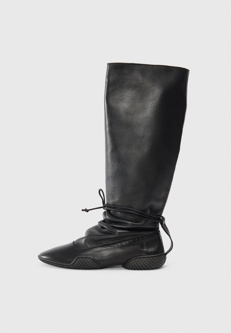 Μαύρα δερμάτινα μποτάκια μέχρι το γόνατο με φαρδύ, λείο άξονα, μυτερή μύτη και υφή στη σόλα. Διαθέτουν λεπτομέρεια με κορδόνι στον αστράγαλο.