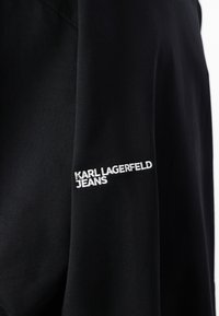 Un material textil de hanorac negru cu text brodat "KARL LAGERFELD JEANS" în alb pe mâneca stângă.