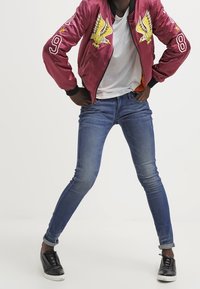 Burgundská bomber bunda s žlutou výšivkou orla a číslem 9, kombinovaná s modrými skinny džíny a černými teniskami.