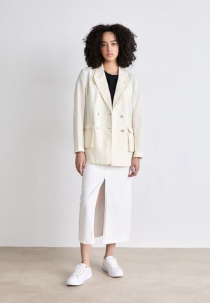 Crème dubbelzijdige blazer met twee voorzakken, gedragen over een zwarte top, gecombineerd met een witte rok met een splits en witte sneakers.