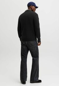 Mann in einem schwarzen Strickpullover, lockeren dunkelgrauen Jeans, schwarzen Schuhen und einer blauen Mütze, der steht und zur Seite schaut, vor einem schlichten Hintergrund.