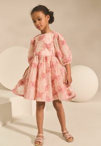 Vestido de tela rosa claro con adornos de rosas, mangas abullonadas y falda acampanada. Combinado con sandalias beige, cuenta con detalles texturizados.