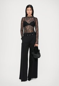 Top din dantelă neagră cu mâneci lungi, suprapus peste un bralette negru, purtat cu pantaloni largi negri, dotati cu buzunare laterale și o geantă structurată.