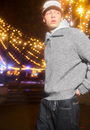 Šedý pletený svetr s vysokým límcem, kombinovaný s tmavými džínovými kalhotami se zřetelným zipem a stříbrnou klíčenkou.