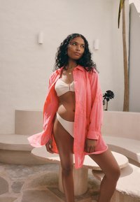 Rožinės spalvos marškiniai su sagomis ir nėrinių detalėmis, nešioti virš tekstūruotos kremo spalvos bikinio. Minkšta medžiaga, laisvas kirpimas ir apykaklės dizainas suteikia išvaizdai išskirtinumo.