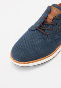 Zapatilla de deportiva azul marino con parte superior de tela texturizada, detalles en cuero marrón, cordones redondos y una suela de goma blanca con un ribete marrón.