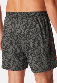 Shorts de bain gris avec un motif de feuilles noires, une taille élastique et une coupe décontractée. Le tissu semble léger et doux.