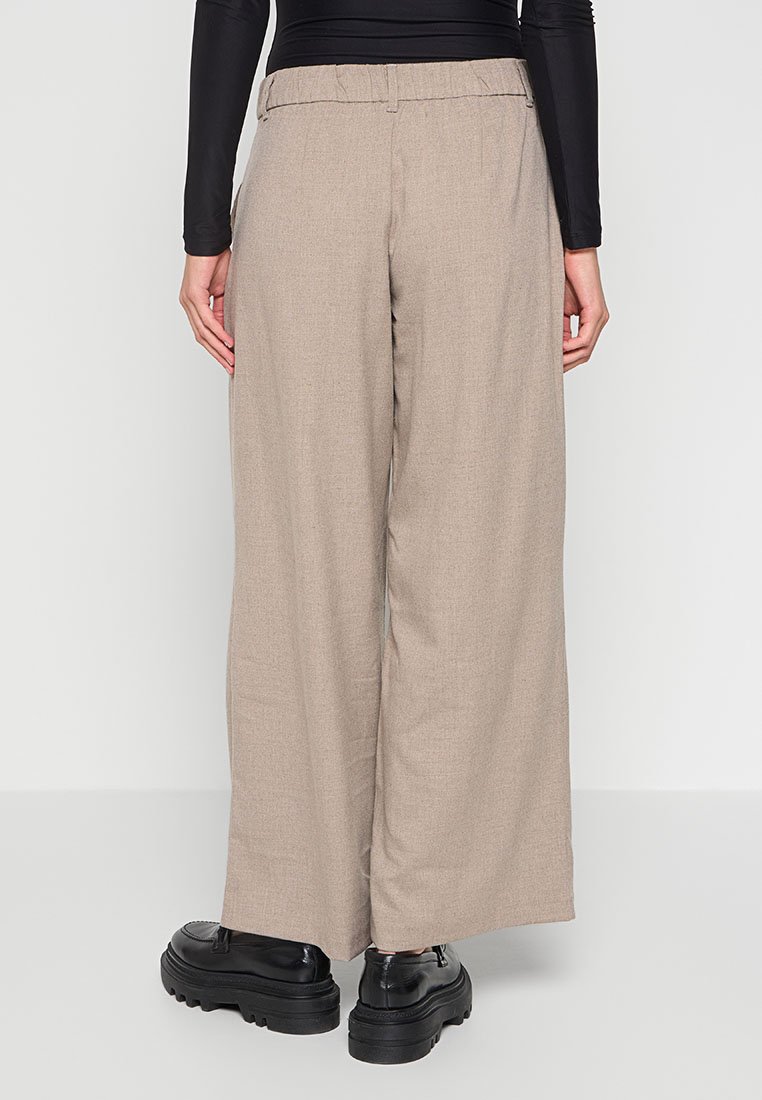 Person in weiten beigen Hosen, schwarzem langärmligen Oberteil und klobigen schwarzen Schuhen, die mit dem Rücken zu einer schlichten Hintergrund stehen.