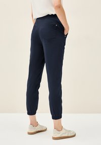 Pantalon de jogging cropped bleu marine avec ceinture élastique, deux poches arrière et poignets côtelés. Associé à des baskets de couleur claire.
