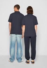 Dva lidé stojí zády k sobě, oba oblečeni v tmavě modrých polo tričkách. Jeden má na sobě světle modré široké džíny, druhý tmavé joggers kalhoty.