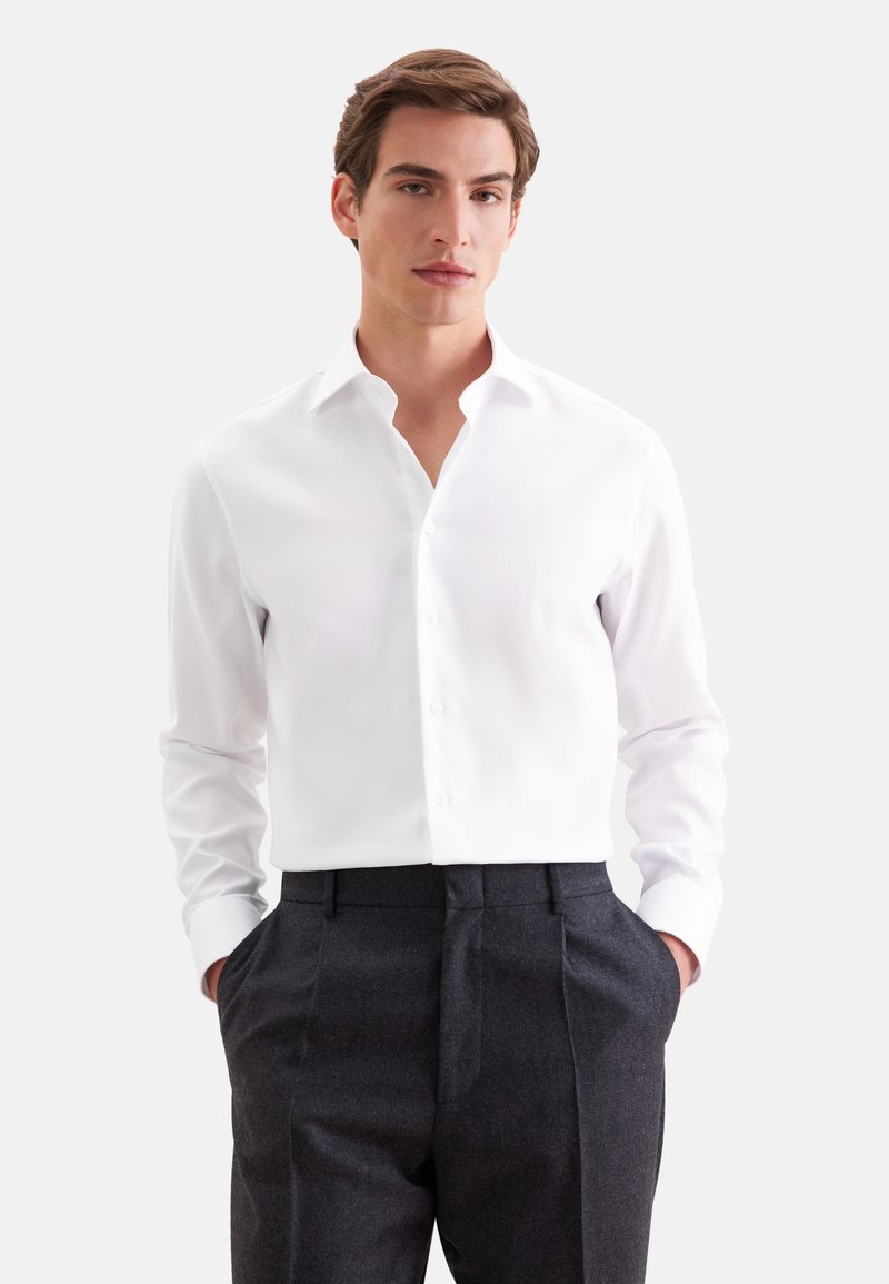 Jeune homme aux cheveux bruns courts portant une chemise blanche à boutons et un pantalon gris foncé, debout les mains dans les poches.