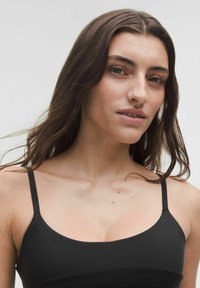 Tânără cu păr lung și castaniu, îmbrăcată cu un top negru cu bretele subțiri, privindu-l pe fotograful din față, pe un fundal simplu și deschis la culoare.