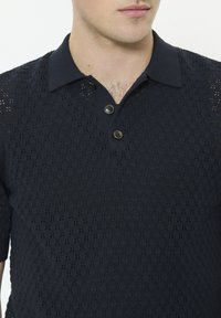 Polo en maille bleu marine avec un motif texturé, doté d'une patte de boutonnage à trois boutons et de manches courtes. Le tissu présente un dessin semblable à de la dentelle.