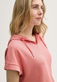 Rosa Kapuzenpullover aus strukturiertem Stoff, mit kurzen Ärmeln und V-Ausschnitt. Mit Kordelzug und silbernen Aglets, vor einem neutralen Hintergrund präsentiert.