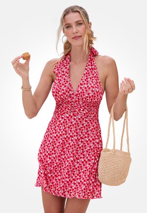 Frau trägt ein rotes Neckholderkleid mit Kirschmuster, hält eine kleine beige gewebte Tasche und ein mundgerechtes Gebäck in einer Hand.