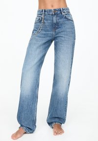 Ventre découvert et pieds visibles, portant un jean bleu taille haute à coupe ample avec un accessoire chaîne argentée sur la poche gauche.