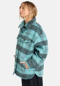 Flanellen jack in turquoise met een grijs plaidpatroon. Heeft een kraag, knoopsluiting en voorzakken. Zacht, gestructureerd materiaal.