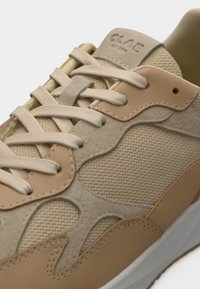 Gros plan d’une basket beige et beige clair avec tissu texturé, accents en daim, lacets arrondis et marquage sur la languette.
