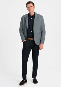 Ljusgrå texturerad kavaj över en marinblå skjorta, kombinerad med mörka jeans och svarta skor. Funktioner inkluderar en klassisk slagkrage och en skräddarsydd passform.