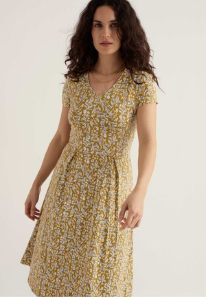 Mujer con cabello rizado y oscuro, vestida con un vestido amarillo mostaza de flores, de mangas cortas y escote en V, de pie frente a un fondo claro y liso.