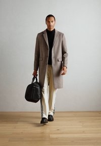 Abrigo de lana marrón claro, cuello alto negro, pantalones crema con franjas laterales y bolso de cuero negro. Textura suave, diseño aerodinámico.