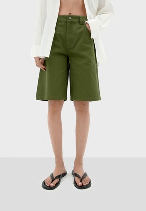 Olijfgroene katoenen bermuda shorts met een rechte snit, een haaksluiting aan de voorkant en zijzakken, in combinatie met zwarte teenslippers.