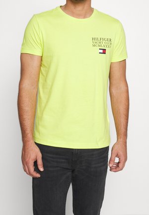 Jasnożółta bawełniana koszulka z okrągłym kołnierzem, z czarnym nadrukiem logo i małym kolorowym detalem w postaci flagi na piersi.