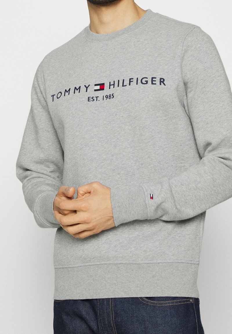 Homme portant un sweat-shirt gris clair Tommy Hilfiger avec logo et texte "EST. 1985", associé à un jean bleu foncé.