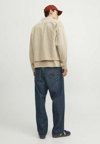Chaqueta de mezclilla beige sobre una camisa de color claro, combinada con unos vaqueros azules holgados. El atuendo se complementa con una gorra roja y zapatos oscuros.