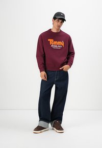 Joven vestido con un suéter burdeos de Tommy Jeans, jeans oscuros de pierna ancha con grandes dobladillos, zapatillas marrones y una gorra oscura contra un fondo blanco.