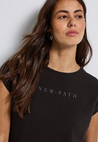 Zwarte t-shirt met ronde halslijn en korte mouwen; heeft de tekst "NEW · PATH" in een subtiele metallic print op de voorkant. Glad textuur.