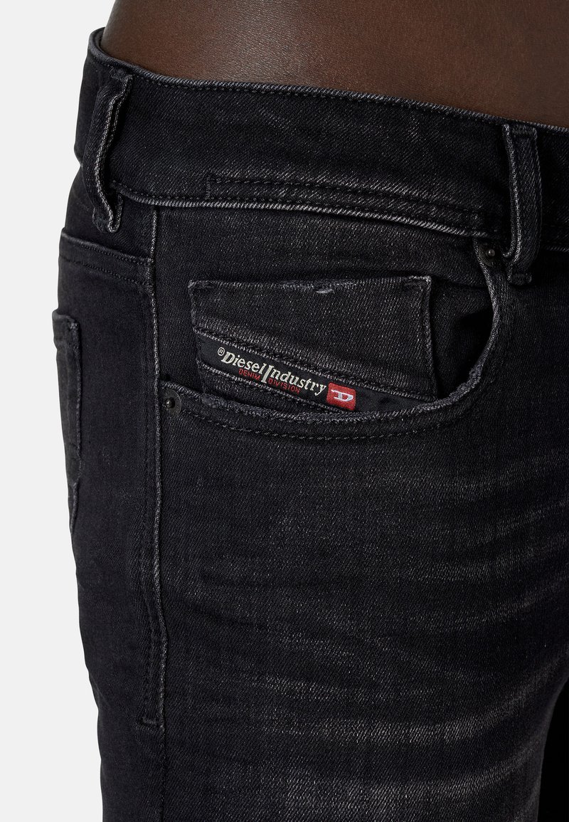 Jean en denim noir avec une taille mi-haute, un léger délavage et un label rouge et blanc sur la poche arrière. Texture lisse et design ajusté.