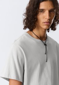 Tricou din bumbac gri deschis, cu manga scurte, avand un decolteu rotund si un detaliu din sfoara neagră legat în față pentru ajustare.