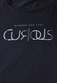 Mörkblå t-shirt med texten "WANDER AND STAY" ovanför ordet "CURIOUS," framhävt med en metallisk glans.