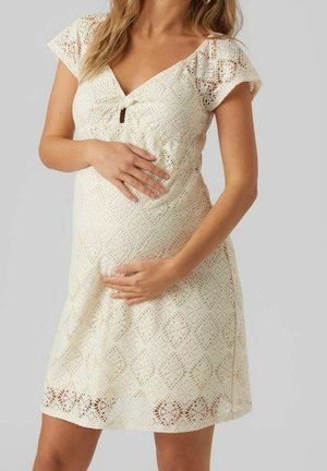 Krajkové těhotenské šaty v krémové barvě s výstřihem do V, krátkými rukávy, nabraným živůtkem a složitým diamantovým vzorem. V oválném výřezu uprostřed.