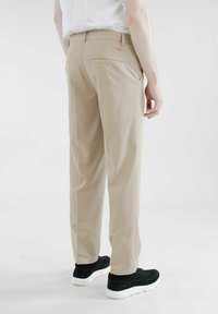 Beige Hosen mit geradem Bein, ausgestattet mit Gesäßtaschen und einem orangefarbenen Logo-Akzent. Getragen mit schwarzen Sneakers vor einem schlichten Hintergrund.