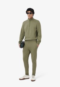 Olijfgroene, lichte katoenen ritsvest met hoge kraag, gecombineerd met bijpassende joggers. Accessoires zijn een zwarte tas en witte sneakers.