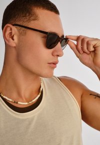 Νεαρός άνδρας ρυθμίζει τα σκουρόχρωμα γυαλιά ηλίου, φορώντας αμάνικη μπεζ μπλούζα και κολιέ με χάντρες, με ένα μικρό τατουάζ στο πάνω μέρος του μπράτσου.