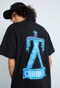 Camiseta de algodón negra con un gráfico de un figura de patrón de circuitos azules en la parte de atrás, con el texto "Carhartt" debajo. Manga corta.